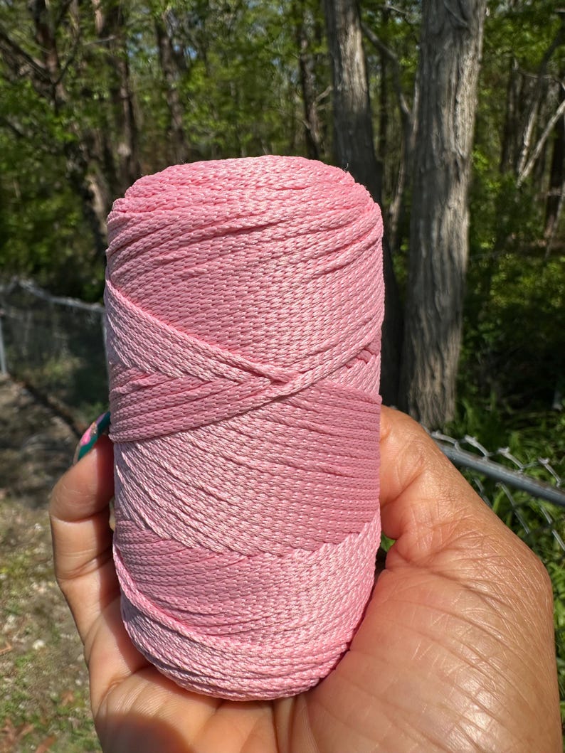 May include: A spool of pink yarn, held in a hand, with varying shades of pink. The yarn is tightly wound, showing a textured surface. The background features green trees and a chain-link fence.