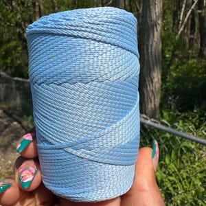 May include: A spool of light blue thread, tightly wound, with a gradient effect from a darker to a lighter shade. The thread is held in a hand with painted nails, set against a blurred natural background.
