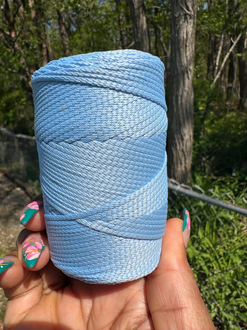May include: A spool of light blue thread, tightly wound, with a gradient effect from a darker to a lighter shade. The thread is held in a hand with painted nails, set against a blurred natural background.