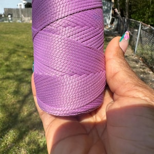 May include: A spool of lavender-colored nylon cord held in a hand. The cord is tightly wound, showing a woven texture. The background includes green grass, a blue sky, and a house.