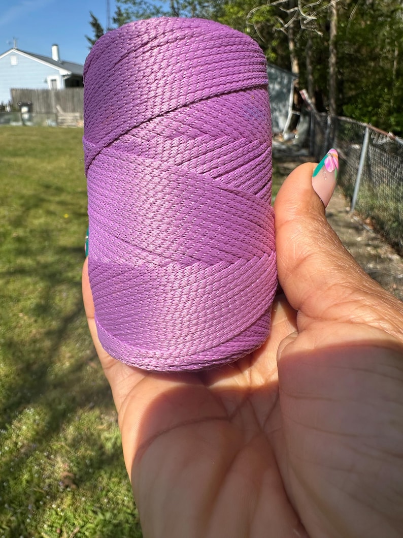 May include: A spool of lavender-colored nylon cord held in a hand. The cord is tightly wound, showing a woven texture. The background includes green grass, a blue sky, and a house.