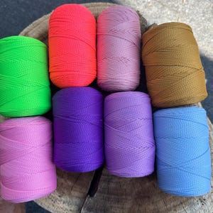 May include: A collection of eight spools of colorful nylon cord in various shades, including green, orange, pink, brown, purple, light purple, and blue. The spools are arranged on a wooden surface, showcasing the vibrant colors and textures of the cord.