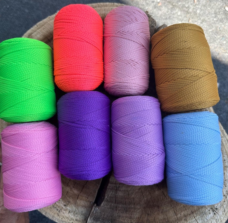 May include: A collection of eight spools of colorful nylon cord in various shades, including green, orange, pink, brown, purple, light purple, and blue. The spools are arranged on a wooden surface, showcasing the vibrant colors and textures of the cord.
