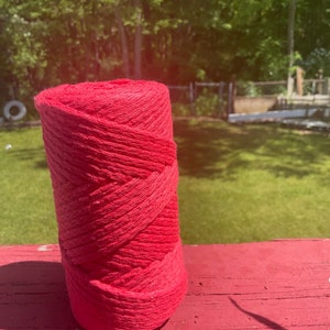 May include: A roll of bright red yarn, likely cotton, is sitting on a red wooden surface. The yarn is wound tightly and has a rough texture.