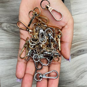 May include: A pile of gold and silver swivel clasps, also known as lobster clasps, in a hand. These clasps are commonly used for attaching straps, chains, or other accessories to bags, purses, or other items.