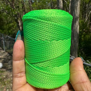 May include: A vibrant green spool of thread, held in a hand. The thread is tightly wound, with varying shades of green creating a textured appearance. The background features lush green foliage and trees.