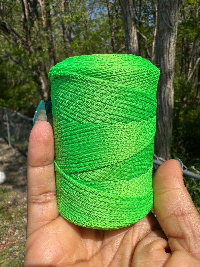 May include: A vibrant green spool of thread, held in a hand. The thread is tightly wound, with varying shades of green creating a textured appearance. The background features lush green foliage and trees.