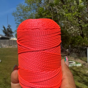 May include: A vibrant, cylindrical spool of red-orange yarn. The yarn is tightly wound, with visible horizontal lines. The image is taken outdoors, with a blurred background of green trees and a blue sky. The yarn is held in a hand.