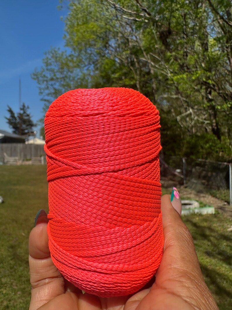 May include: A vibrant, cylindrical spool of red-orange yarn. The yarn is tightly wound, with visible horizontal lines. The image is taken outdoors, with a blurred background of green trees and a blue sky. The yarn is held in a hand.