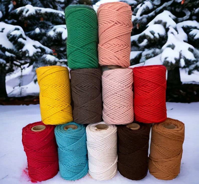 May include: A collection of twelve cylindrical spools of macrame cord arranged in a pyramid stack. The spools feature a variety of solid colors including green, peach, yellow, brown, light pink, red, teal, cream, dark brown, and tan.