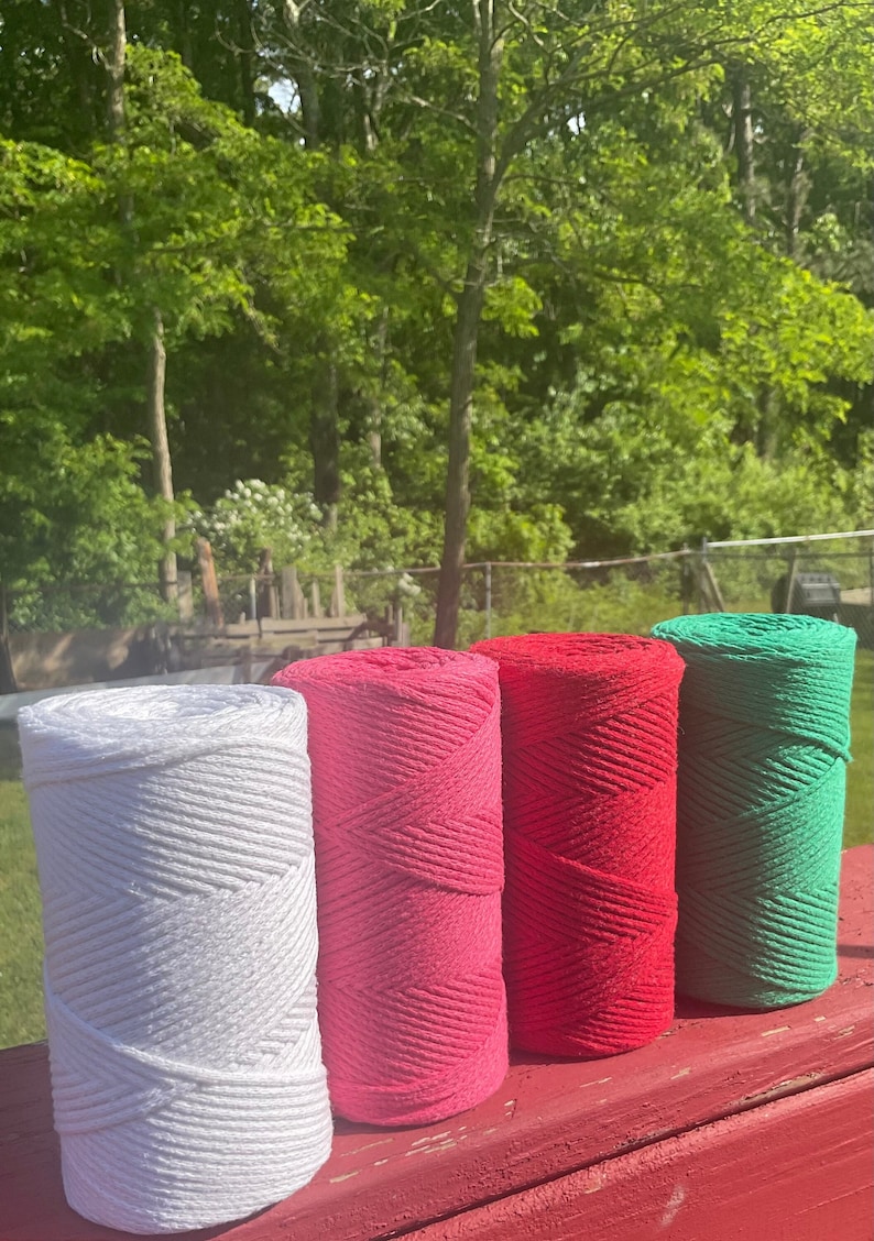May include: Four spools of cotton rope in white, pink, red, and green. The spools are arranged in a row on a red wooden surface.