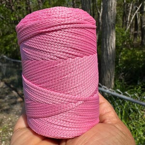 May include: A roll of pink cord held in a hand. The cord is a gradient of pink shades, from light to dark. The cord appears to be tightly wound and has a textured surface. The background is a blurred view of trees and foliage.