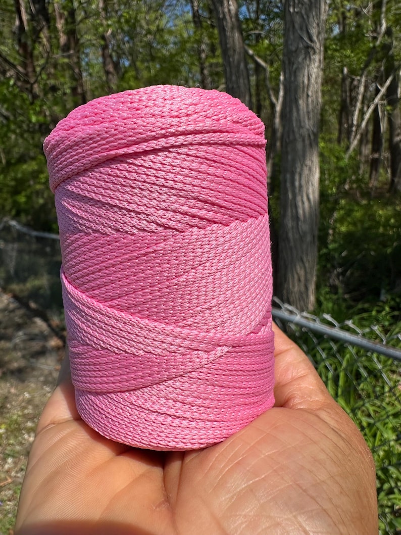 May include: A roll of pink cord held in a hand. The cord is a gradient of pink shades, from light to dark. The cord appears to be tightly wound and has a textured surface. The background is a blurred view of trees and foliage.
