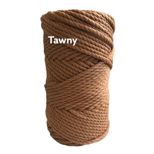 May include: A cylindrical spool of thick, tawny brown macrame cord wound in a tight, continuous spiral pattern. The word Tawny is printed in white sans-serif text on the upper left side of the cord spool against the textured brown surface.