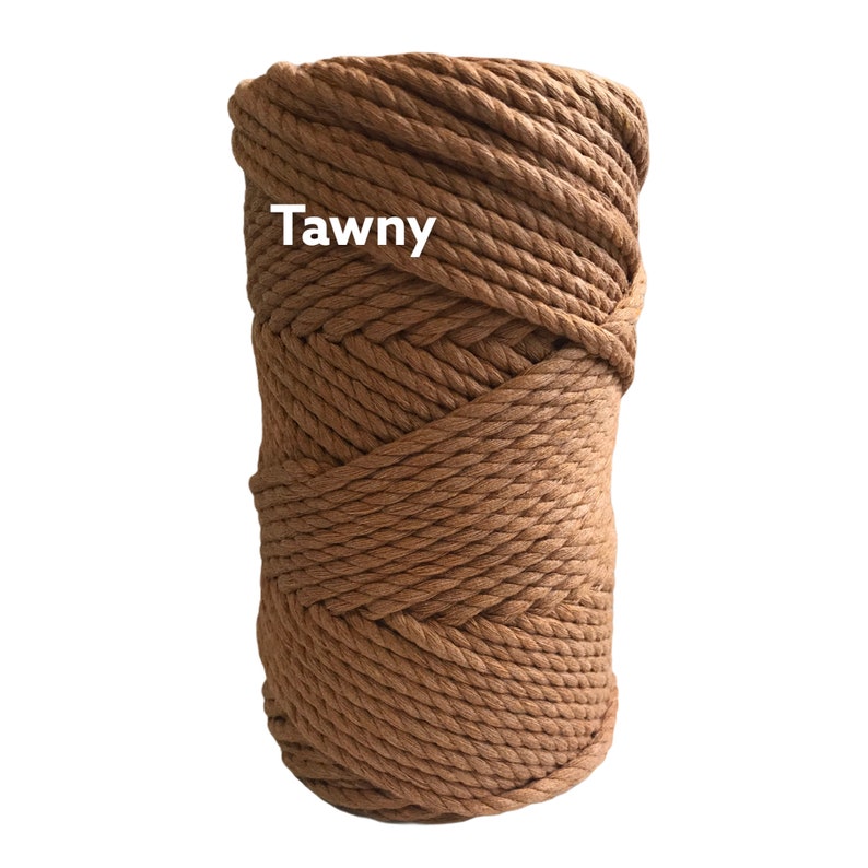May include: A cylindrical spool of thick, tawny brown macrame cord wound in a tight, continuous spiral pattern. The word Tawny is printed in white sans-serif text on the upper left side of the cord spool against the textured brown surface.