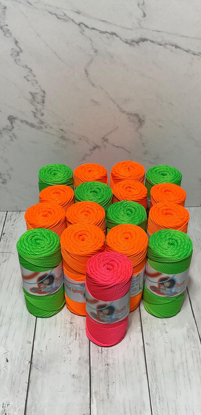 May include: A collection of neon green, orange, and pink spools of string. The spools are arranged in a pyramid shape and have labels that say "Soft n' Strong" and "String Yarn".