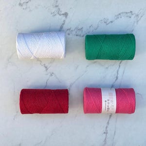 May include: Four spools of cotton cord in different colors: white, green, red, and pink. The spools are arranged in a square pattern on a white marble surface.