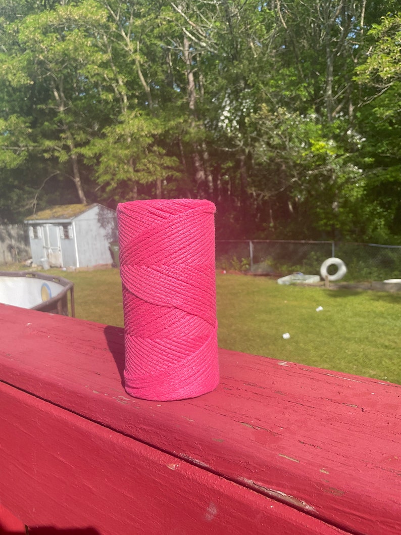 May include: A pink spool of twine, sitting on a red wooden surface. The spool is made of natural fibers and has a rough texture.