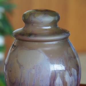 Puede incluir: Un tarro de cerámica con cuerpo redondo y tapa. El tarro tiene un esmalte marrón claro con vetas blancas y moradas.