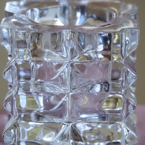 May include: A clear glass candle holder with a geometric, textured design. The candle holder is square-shaped with a cylindrical opening at the top. It is made of stacked, square blocks. The glass reflects light, creating a sparkling effect.