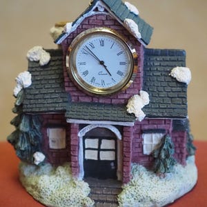 May include: A small, red brick building with a white clock face and black Roman numeral hour markers. The building has a snow-covered roof and a black door with a white window. The clock face reads "Quartz".