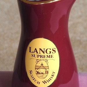 Peut inclure: Pichet en céramique de couleur bordeaux avec un bord doré. Le pichet porte une étiquette ovale avec les mots "LANGS SUPREME SCOTCH WHISKY" et une illustration d'un bâtiment. Objet décoratif.