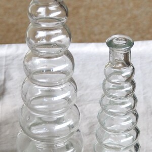 May include: Two clear glass bottles with a stacked bubble design. One bottle is taller than the other. Cork stoppers are at the base of each bottle. The bottles are on a white surface.