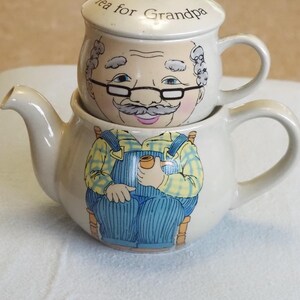 May include: A cream-coloured ceramic teapot and teacup set. The teacup, with a handle, is stacked on top of the teapot. The teapot features a cartoon of a man in overalls holding a pipe. The teacup lid reads "Tea for Grandpa".