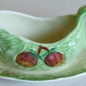 Puede incluir: Una salsera de cerámica verde con asa y un plato a juego. La salsera está decorada con un diseño de hojas y dos frutas rojas y amarillas.
