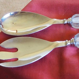 Puede incluir: Un par de cubiertos de ensalada plateados con mangos transparentes en forma de esfera. La cuchara y el tenedor descansan sobre una superficie roja oscura. La cuchara tiene un cuenco ancho y ovalado, y el tenedor tiene tres púas.