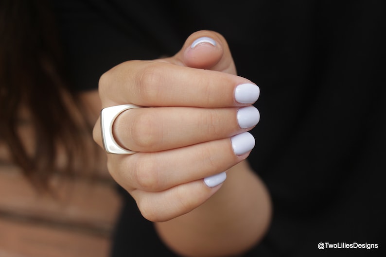 May include: A silver ring with a square face and a curved inner band. The ring is worn on a hand with light purple nail polish. The background is black.