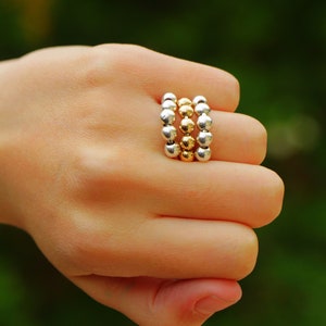 Könnte beinhalten: Eine Hand trägt drei übereinanderliegende Ringe. Die Ringe bestehen aus silbernen und goldenen Perlen.