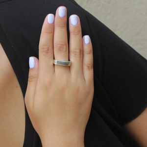 May include: A silver rectangular ring is worn on a hand with light purple nail polish. The hand is resting on a black garment. The background is a neutral beige color.