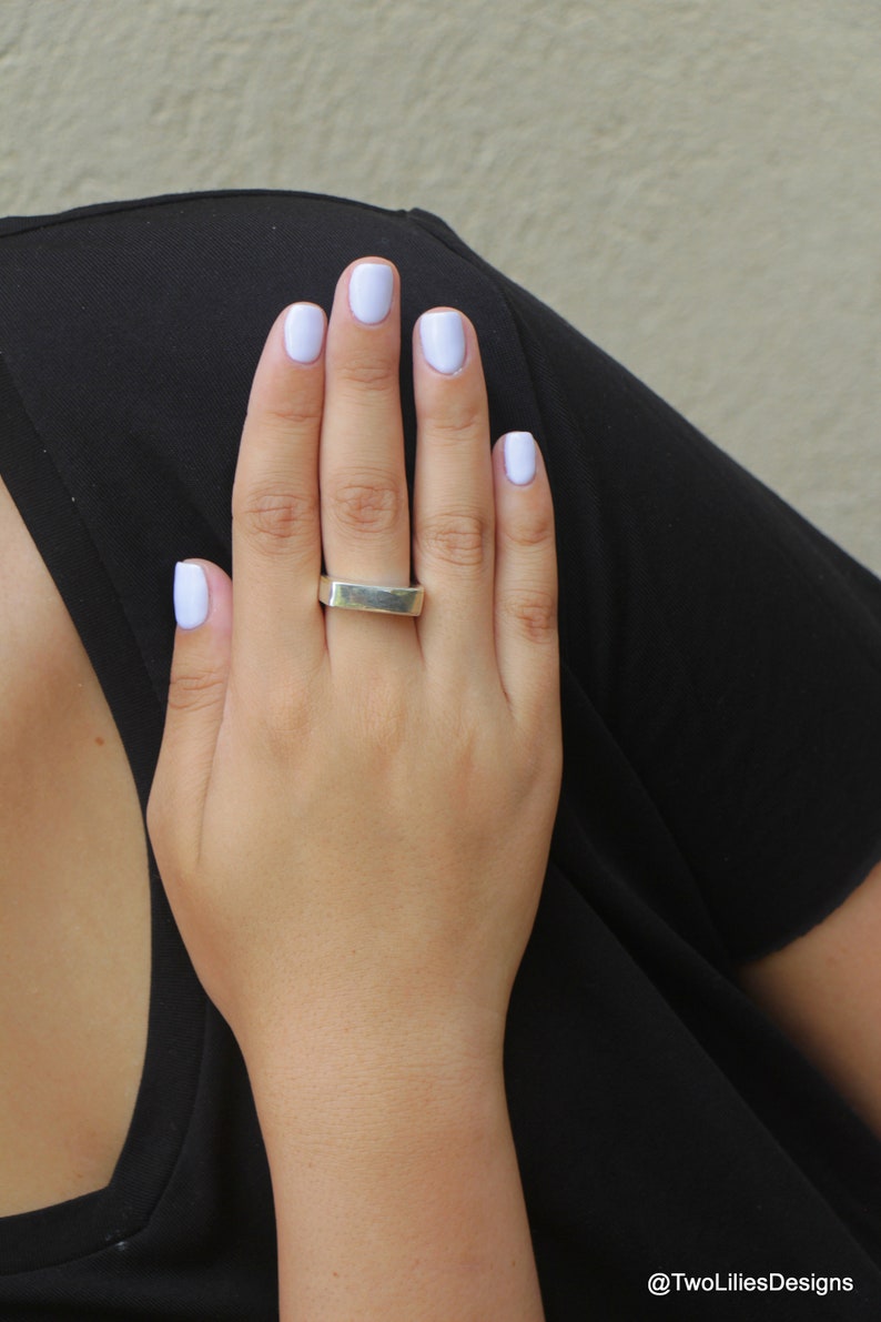 May include: A silver rectangular ring is worn on a hand with light purple nail polish. The hand is resting on a black garment. The background is a neutral beige color.
