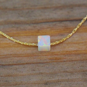 Puede incluir: Un delicado collar de cadena dorada con un solo colgante de ópalo de forma cuadrada. El ópalo es iridiscente, con tonos de blanco, rosa y azul.