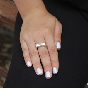May include: A silver ring with a rectangular design is worn on a finger. The hand has white nail polish and rests on black fabric. The ring is the focal point, showcasing a minimalist style.