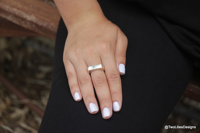 May include: A silver ring with a rectangular design is worn on a finger. The hand has white nail polish and rests on black fabric. The ring is the focal point, showcasing a minimalist style.