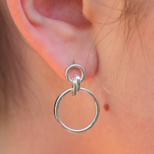 Pode incluir: Brincos de prata com dois círculos e um detalhe de nó. Os brincos são usados no lóbulo da orelha.