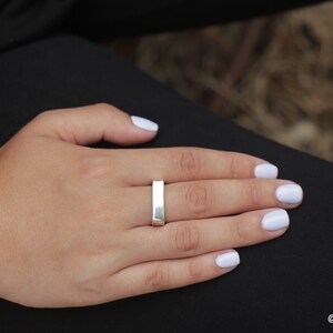 May include: A silver rectangular ring is worn on a person's finger. The ring is a simple, minimalist design. The person's hand is resting on a black surface, with white nail polish. The background is blurred.