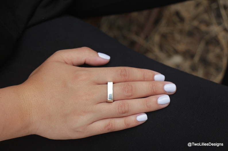 May include: A silver rectangular ring is worn on a person's finger. The ring is a simple, minimalist design. The person's hand is resting on a black surface, with white nail polish. The background is blurred.
