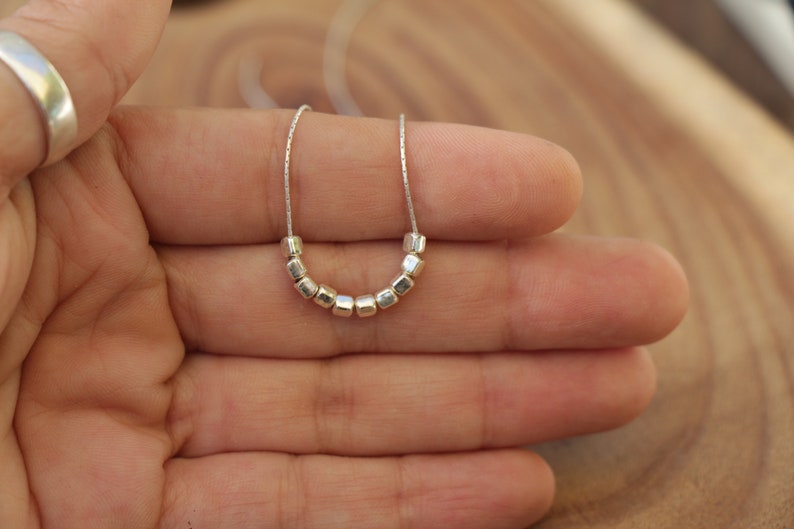 Op de afbeelding: Een zilveren ketting met een ketting en een rij kleine, vierkante kralen.