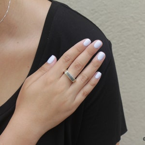 May include: A silver-colored ring with a rectangular design. The ring is worn on a finger with light blue nail polish. A silver necklace is also visible. The person is wearing a black shirt.