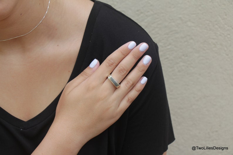 May include: A silver-colored ring with a rectangular design. The ring is worn on a finger with light blue nail polish. A silver necklace is also visible. The person is wearing a black shirt.