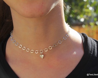 May include: A silver chain choker necklace with a small heart pendant. The necklace is worn by a person with a black shirt.