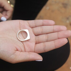 May include: A silver ring with a square face and a rounded band is held in an open hand. The ring is smooth and shiny. The background is blurred, with a hint of a person's clothing and a natural surface.