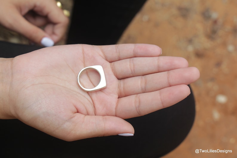 May include: A silver ring with a square face and a rounded band is held in an open hand. The ring is smooth and shiny. The background is blurred, with a hint of a person's clothing and a natural surface.