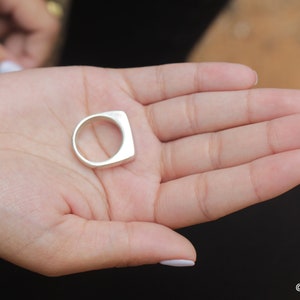 May include: A silver ring with a square face and a rounded band is held in an open hand. The ring has a minimalist design and a polished finish. The ring is a piece of jewelry.