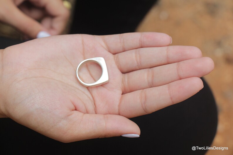 May include: A silver ring with a square face and a rounded band is held in an open hand. The ring has a minimalist design and a polished finish. The ring is a piece of jewelry.