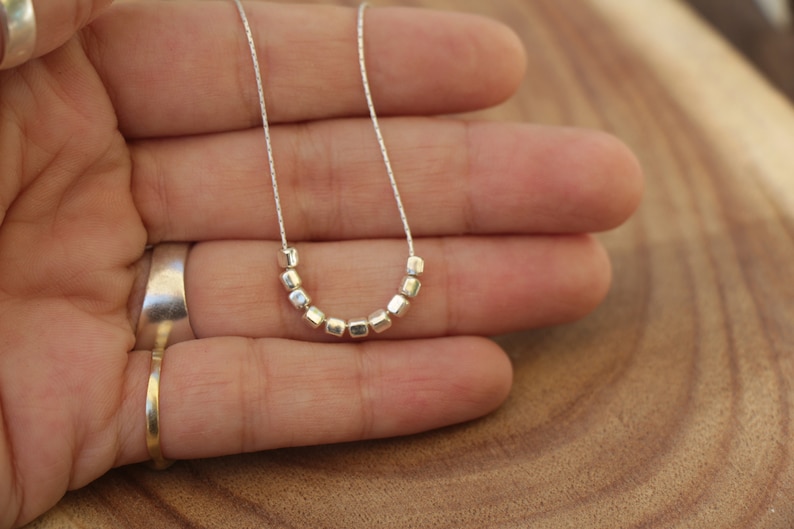 Op de afbeelding: Een zilveren ketting met een ketting en een rij kleine, vierkante kralen.