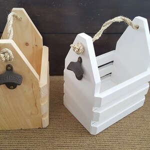 May include: Two wooden bottle carriers with rope handles and bottle openers. One carrier is natural wood and the other is painted white.